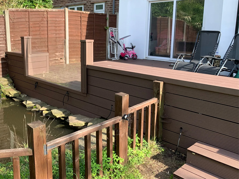 side view garden fence surround ,raised sitting area, decking brown composite boards and glass balustrade near fish pound