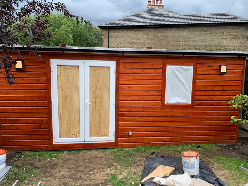 garden room front side view, external walls cover in timber cladding painted in golden cedar, white UPVC windows and double French doors 